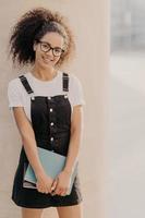 toma vertical de un alegre joven estudiante afro que lleva blocs de notas o diario, usa camiseta blanca, sarafan negro, listo para tener clases, se para al aire libre. personas, estudio y concepto universitario. foto