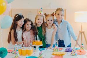 Friendly children embrace while pose near festive table, blow candles on cake, have party mood, celebrate birthday or special occasion, have joyful expressions. Childhood, fun and entertainment photo
