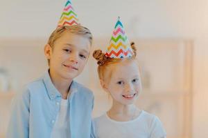 Horizontal shot of happy girl and boy wears cone party hats, celebrate birthday together, have good mood, wait for guests, pose indoor against blurred background. Children, holiday, celebration photo