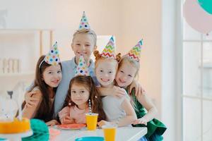 Photo of happy friends gather around table, wear party hats, embrace and look gladfully at camera, smile positively, celebrate birthday at home together, have fun