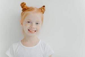 Positive attractive little girl with double ginger buns, freckles on face, toothy smile, dressed in casual t shirt, stands against white background, empty space for your advert. Children concept photo