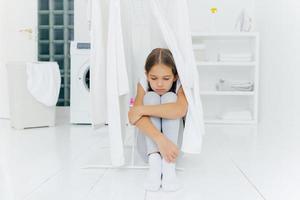 Small adorable girl sits on floor, being punished by parents, poses near clothes dryer, focused down with sad expression, washing machine, basket with laundry and console, thinks over something photo