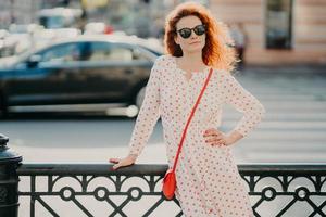 toma al aire libre de una mujer pelirroja satisfecha mantiene una mano en la cintura, otra en la calle, por lo tanto, posa sobre un entorno urbano borroso, usa gafas de sol y vestido, tiene manicura roja. copie el espacio a un lado para el texto foto