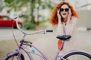 Happy woman has curly foxy hair, leans at saddle of her bicycle, has outdoor promenade during sunny summer day, wears stylish shades and dress, poses over urban setting. Healthy lifestyle and rest