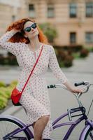 Healthy outdoor activity concept. Lovely curly red haired woman with dreamy expression, uses bicycle for traveling across city, wears fashionable white dress and sunglasses, poses in urban setting photo