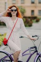 Street style and active rest concept. Curly red haired woman keeps hand on frame of sunglasses, wears white dress, focused into distance, waits for friend, going to have outdoor trip on bicycle photo