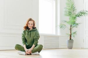 Beautiful satisfied slim woman with ginger hair, appealing appearance, sits crossed legs, wears green sweatsuit, keeps perfect body shape, stays fit. Athletic training and healthy lifestyle concept photo