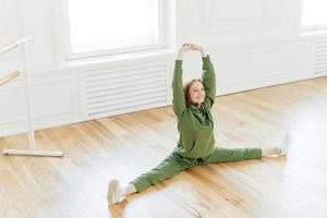 Strong flexible curly red haired woman does side leg split, raises hands, wants to have perfect body, wears sportsclothes, has training in ballet studio. Talented dancer has rehearsal indoor photo