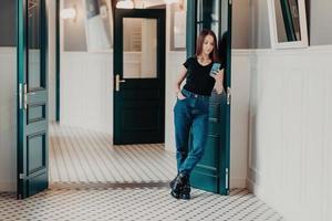 Full length shot of serious fashionable woman concentrated into cell phone, wears black t shirt, jeans and boots, reads information in internet, stands indoor near doors connected to wireless internet photo