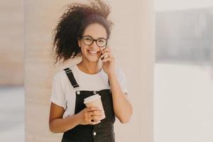 Pleasant looking curly teenage girl wears transparent glasses, talks on mobile phone, has coffee break, dressed in white t shirt and overalls, enjoys spare time and conversation. People and technology photo