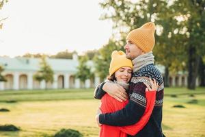 People and closeness concept. Young couple in love have date, embrace each other, feel support, being alone in park, have perfect relationships. Handsome man in warm sweater and hat hugs girlfriend photo