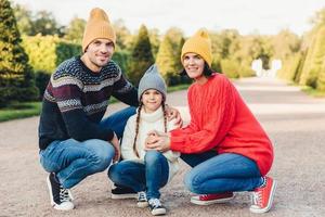 Friendly family wear knitted clothes, have walk together, admire splendid autumn weather. Affectionate young parents and their little cute daughter play together outdoor. Relationship concept photo