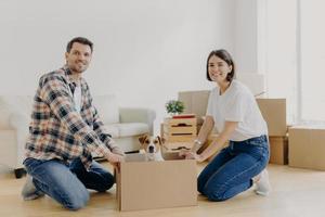 Positive happy husband and wife stand on knees near carton box with cute dog, enjoy relocation in new apartment, smiles pleasantly at camera. Pedigree pet rides in cardboard container on floor photo