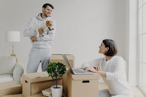 Happy family couple move into new flat for living. Wife works on laptop computer, searches information in internet, makes shopping online, man stands with dog, unpacks belongings from boxes. photo