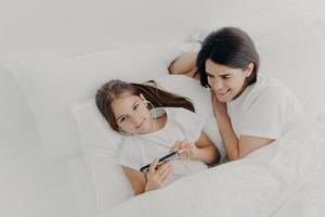Caring mom looks attentively at her little daughter who wakes up early, listens music in earphones, searches new song in playlist of modern smartphone, lie under soft white blanket. Top view photo