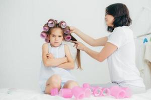 Displeased child keeps hands crossed, looks with sullen expression, being discontent as her mother winds curlers on her hair, doesnt want to have curly hairstyle, pose against white background photo