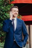 Vertical shot of satisfied happy male employee has telephone conversation, dressed in black suit, keeps hand in pocket, has glad expression, gentle smile on face, being confident in everything photo