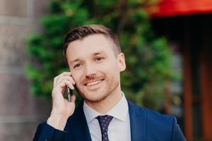 Successful male office worker has conversation about business affairs via cell phone, dressed in formal clothes, going to have lunch break at cafe, poses outside, has positive smile on face. photo