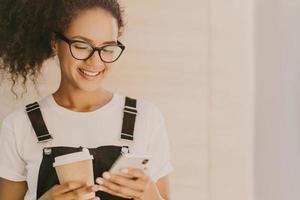 adorable teeange girl con cabello rizado, revisa las noticias, mensajes en las redes sociales, usa conexión gratuita a Internet, usa anteojos ópticos, camiseta blanca informal y overoles, bebe café para llevar foto