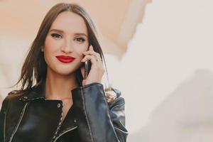 Attractive lovely female enjoys tariffs in roaming, talks on mobile phone with best friend, dressed in fashionable clothes, wears red lipstick, makeup, stands on blurred background of some building photo