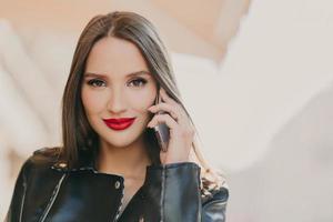Close up shot of lovely woman talks with friend via mobile app, has makeup and red lips, dressed in leather jacket, has spare time outdoor. Online communication concept. Beauty and feminity. photo