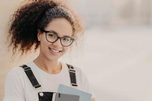 Horizontal shot of beautiful woman with crisp hair combed in pony tail, carries notepad, wears white t shirt and overalls, enjoys studying, poses against blurred background with empty space for text photo