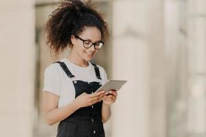 Pretty curly haired girl looks at digital tablet screen with smile, reads some text or electronic book, wears spectacles for vision correction, white t shirt and dungarees, enjoys high speed internet photo