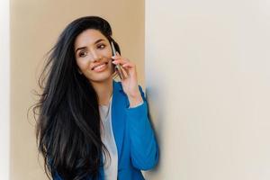 Happy young female student talks via cell phone after exam, shares her success. Brunette woman entrepreneur enjoys phone conversation, looks aside gladfully, wears formal outfit, communicates photo