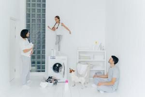 Happy family do laundry at home, father sits on floor in lotus pose, mother stands with white towel, look at child who dances happily on washing machine, pedigree dog near. Household chores. photo