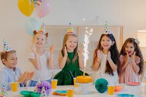 Friends on birthday party, look gladfully at cake, stand near festive table, wear cone hats, clap hands, play together, pose in decorated room. Childhood and holiday concept