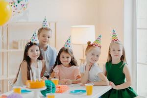 Children, celebration and birthday concept. Positive children have fun together at party, wear cone hats, eat delicious cake, play interesting games, being in good mood photo