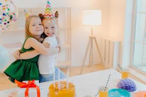 Friendly two girls embrace and have good relationship, stand near festive table with cake, celebrate birthday together, stand in living room. Glad female sisters enjoy holiday, special occasion photo