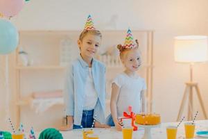 Indoor shot of small girl and boy wear party hats, stand near festive table with cake, paper cups and present, celebrate birthday together, pose in white spacious room. Celebration concept. photo