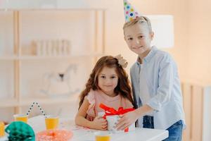 una niña y un niño felices sostienen una caja de regalo con una cinta roja, preparan una sorpresa en el cumpleaños, posan en una mesa blanca, usan sombreros de fiesta de cono, tienen expresiones positivas. concepto de infancia foto