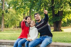 Father and mother sit between their daughter against trees or nature background, show something to her, indicate with fore fingers. Smiling parents show bird to little beautiful girl. Parenthood photo