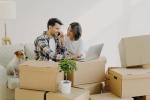 Happy husband and wife move in new bought house, buy furniture online, use modern technology, man surfs internet via laptop and talk over phone, sits on sofa with girlfriend and dog, many carton boxes photo