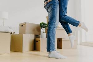 Faceless young couple move in new home, man lifts woman, have fun, surrounded with unpacked cardboard boxes, start new life in recently bought abode. Family, relocation and moving day concept photo