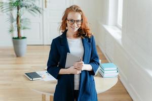 Happy good looking female office worker enjoys online banking service, checks balance on touch pad, reads financial news, wears elegant clothes, stands near table in cozy cabinet, smiles gently photo