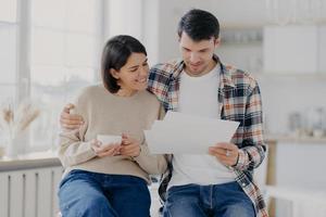 Caring man embraces his wife, look through documents with happy expressions, study payment bills, drinks coffee, dressed in casual wear, pose in spacious room with blurred background. Family and work photo