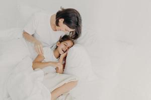 Overjoyed mum and daughter have fun together in bedroom, pose under soft blanket. Happy small girl spends leisure time with mother, has toothy smile, expresses happiness. Children and parents photo