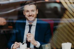 Horizontal shot of happy businessman dressed in formal clothing, drinks aromatic hot beverage, looks at window while sits in cafeteria, has pleased expression. People, rest and lifestyle concept photo