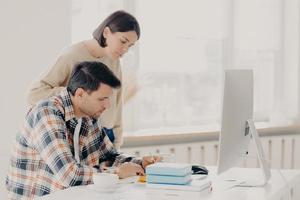 Husband and wife work on paper report together, check information, sit in front of computer, drinks coffee, dressed in casual wear, collaborate for teamworking. Family and remote work concept photo