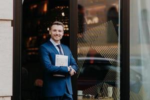 Horizontal shot of handsome male with cheerful expression, dressed in formal suit, holds magazine, stands against cafe background, going to have lunch during work break. Successful businessman