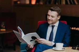 Portrait of happy male comapny owner reads magazine, rests in cozy coffee shop, has happy expression, has break after business meeting, dressed in formal clothes. People, rest and leisure concept photo