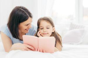 afectuosa madre joven con cabello oscuro, mira felizmente a su pequeña hija, lee un libro interesante o un cuento de hadas juntos, disfruta del ambiente doméstico en casa. gente, familia, concepto de infancia foto