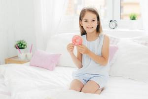 Lovely small girl holds delicious doughnut in hands, going to have breakfast, poses on bed in cozy white bedroom, looks at camera with positive expression. Children, dessert, bed time concept photo