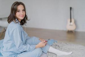 Photo of pleased relaxed woman dressed in casual clothes, sits on comfortable bed, listens music via application on smart phone and earphones, rests in bedroom alone, acoustic guitar in background