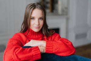 Photo of young good lookng brunette woman enjoys day off at home, has serious look at camera, wears warm loose red sweater, poses at home against blurred background. Coziness and rest concept