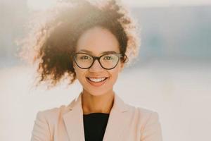 Headshot of cheerful curly woman with positive expression, wears spectacles, white elegant jacket, looks straightly at camera, stands outdoor, has curly hair expresses good emotions. Ethnicity concept photo