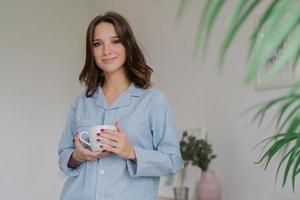 Waist up shot of pleasant looking young woman dressed in striped pyjamas, drinks hot beverage after awakening, has sparre time at home, stands indoor against cozy interior. People and rest concept photo